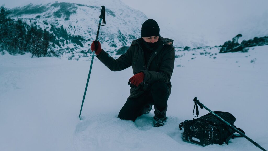 A man kneels in a snowy mountain setting, holding a trekking pole, with winter gear.